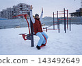 Side view of a bearded man exercising on street fitness equipment at snowy public sports ground in winter. 143492649
