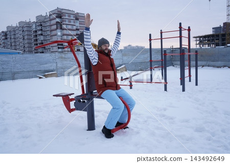 Side view of a bearded man exercising on street fitness equipment at snowy public sports ground in winter. Side view of a bearded man exercising on street fitness equipment at snowy public sports ground in winter. 143492649