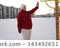 Side view of a mature woman in red jacket lifting arm to warm up at winter public sports ground. 143492651