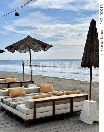 Cushioned beach daybeds face ocean waves beneath woven parasols in soft daylight. Cushioned beach daybeds face ocean waves beneath woven parasols in soft daylight. 143492918
