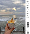 Hand holds an ice cream cup beside ocean waves under soft sunset light. 143493695