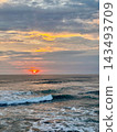 Orange sunset illuminates layered clouds above breaking ocean waves and white foam. 143493709