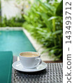White coffee cup rests on wicker furniture beside a tropical pool in soft daylight. 143493712