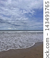 Foamy ocean waves spread across smooth beach sand beneath layered clouds in soft daylight. 143493765