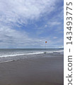 Red and yellow beach flag stands beside breaking waves beneath a softly lit cloudy sky. 143493775