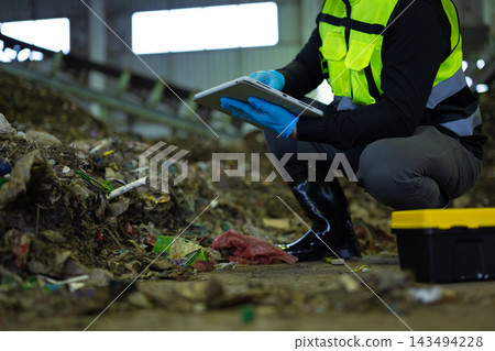 Industrial worker in safety gear crouching by a waste pile, using a digital tablet to monitor recycling and compost operations Industrial worker in safety gear crouching by a waste pile, using a digital tablet to monitor recycling and compost operations 143494228