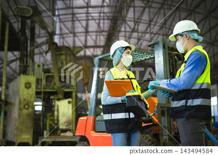 Asian engineers in hardhats discussing logistics and facility maintenance with tablet and clipboard near an industrial forklift Asian engineers in hardhats discussing logistics and facility maintenance with tablet and clipboard near an industrial forklift 143494584