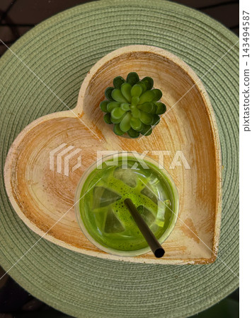 Iced matcha on heart-shaped tray with succulent decoration. Iced matcha with straw sits on heart-shaped tray beside small succulent decor. Overhead view shows ice cubes, circular green woven placemat Iced matcha on heart-shaped tray with succulent decoration. Iced matcha with straw sits on heart-shaped tray beside small succulent decor. Overhead view shows ice cubes, circular green woven placemat 143494587