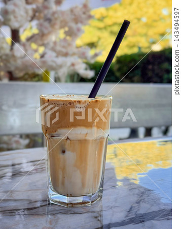 Iced Latte with Straw on Marble Cafe Table. Iced coffee with milk and foamy brown surface fills tall glass with a black straw. Flowers and outdoor seating appear softly blurred behind cafe table 143494597