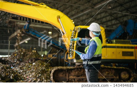 Industrial engineer with a digital tablet oversees waste sorting and recycling operations alongside heavy excavator machinery inside an urban disposal plant. Industrial engineer with a digital tablet oversees waste sorting and recycling operations alongside heavy excavator machinery inside an urban disposal plant. 143494685