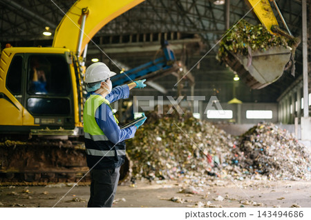Industrial engineer with a digital tablet oversees waste sorting and recycling operations alongside heavy excavator machinery inside an urban disposal plant. Industrial engineer with a digital tablet oversees waste sorting and recycling operations alongside heavy excavator machinery inside an urban disposal plant. 143494686