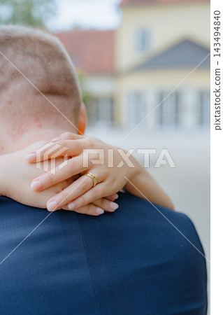 person wearing engagement ring on hand with clasped hands behind back in romantic outdoor setting with blurred buildings, dark clothing and metal ring, gold and dark blue tones, over-the-ey. 143494840