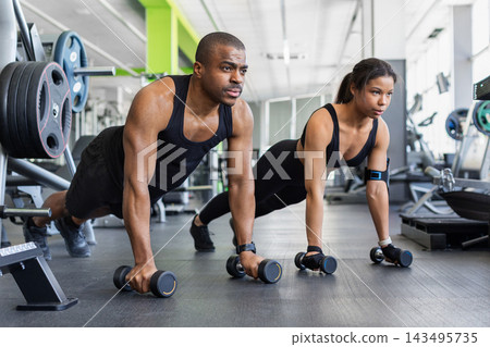Black couple performing high plank exercises while gripping dumbbells on gym floor 143495735
