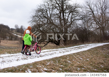 Joyful winter ride through a snowy path with a child in bright green jacket and a parent guiding on a pink bicycle amidst a serene nature backdrop Joyful winter ride through a snowy path with a child in bright green jacket and a parent guiding on a pink bicycle amidst a serene nature backdrop 143497568