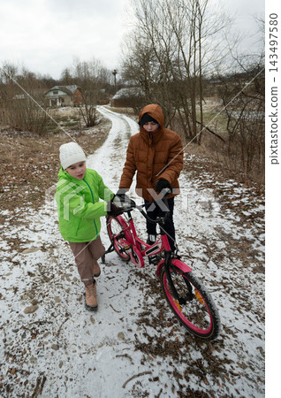 Children enjoying a wintery day as one assists the other with a pink bicycle on a snowy path in a serene rural landscape Children enjoying a wintery day as one assists the other with a pink bicycle on a snowy path in a serene rural landscape 143497580