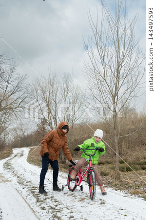 Winter adventure unfolds on a snowy path as a child learns to ride a bicycle with the support of an encouraging adult amidst bare trees and a cloudy sky 143497593