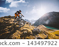 Mountain biker descending a rugged alpine trail above a mountain lake, surrounded by steep peaks and golden grass in warm sunlight, capturing adventure, speed and extreme outdoor sport. 143497922