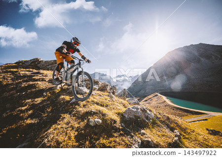 Mountain biker descending a rugged alpine trail above a mountain lake, surrounded by steep peaks and golden grass in warm sunlight, capturing adventure, speed and extreme outdoor sport. Mountain biker descending a rugged alpine trail above a mountain lake, surrounded by steep peaks and golden grass in warm sunlight, capturing adventure, speed and extreme outdoor sport. 143497922