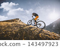 Mountain biker riding across a rugged alpine slope with dramatic mountain scenery, warm sunlight and rocky terrain, capturing outdoor adventure, mountain biking and high altitude freedom. 143497923