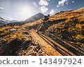 Mountain biker riding down a steep rocky alpine trail with dramatic mountains and a turquoise lake below, captured in warm sunlight during an adventurous high altitude descent ride. 143497924