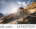 Mountain biker riding down a steep rocky alpine trail with dramatic mountains and a turquoise lake below, captured in warm sunlight during an adventurous high altitude descent ride. 143497926