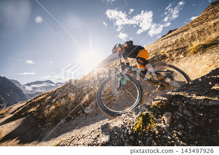 Mountain biker riding down a steep rocky alpine trail with dramatic mountains and a turquoise lake below, captured in warm sunlight during an adventurous high altitude descent ride. Mountain biker riding down a steep rocky alpine trail with dramatic mountains and a turquoise lake below, captured in warm sunlight during an adventurous high altitude descent ride. 143497926