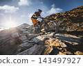 Mountain biker descending a rugged rocky alpine trail in bright sunlight, surrounded by dramatic mountain terrain and blue sky, capturing action, adventure, skill and extreme outdoor sport. 143497927