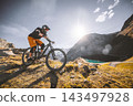 Mountain biker descending a rugged alpine trail above a mountain lake, surrounded by steep peaks and golden grass in warm sunlight, capturing adventure, speed and extreme outdoor sport. 143497928