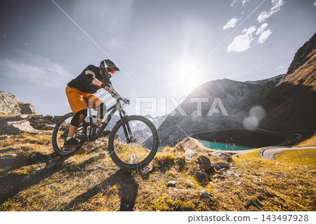 Mountain biker descending a rugged alpine trail above a mountain lake, surrounded by steep peaks and golden grass in warm sunlight, capturing adventure, speed and extreme outdoor sport. Mountain biker descending a rugged alpine trail above a mountain lake, surrounded by steep peaks and golden grass in warm sunlight, capturing adventure, speed and extreme outdoor sport. 143497928