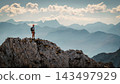 Hiker standing on a rocky summit during a via ferrata in the Austrian Alps near Innsbruck, Tyrol, with dramatic mountain peaks and a vast alpine landscape in the background. 143497929