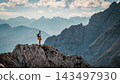 Hiker standing on a rocky summit during a via ferrata in the Austrian Alps near Innsbruck, Tyrol, with dramatic mountain peaks and a vast alpine landscape in the background. 143497930