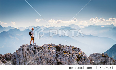 Hiker standing on a rocky summit during a via ferrata in the Austrian Alps near Innsbruck, Tyrol, with dramatic mountain peaks and a vast alpine landscape in the background. Hiker standing on a rocky summit during a via ferrata in the Austrian Alps near Innsbruck, Tyrol, with dramatic mountain peaks and a vast alpine landscape in the background. 143497931