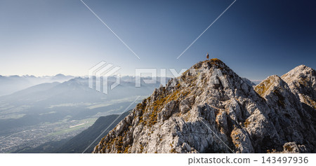 Hiker standing on a rocky summit during a via ferrata in the Austrian Alps near Innsbruck, Tyrol, with dramatic mountain peaks and a vast alpine landscape in the background. Hiker standing on a rocky summit during a via ferrata in the Austrian Alps near Innsbruck, Tyrol, with dramatic mountain peaks and a vast alpine landscape in the background. 143497936