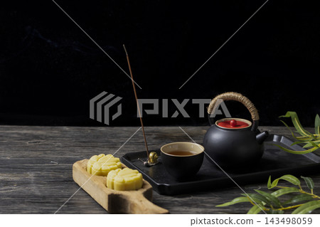 Traditional Chinese tea set with black teapot, cup of tea, burning incense stick, and mung bean cakes on a dark wooden table Traditional Chinese tea set with black teapot, cup of tea, burning incense stick, and mung bean cakes on a dark wooden table 143498059