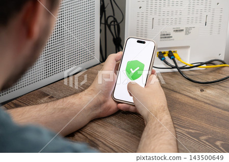 A man holds a smartphone with a green checkmark shield icon on the display to show successful network security settings while sitting at a desk near a white internet router device A man holds a smartphone with a green checkmark shield icon on the display to show successful network security settings while sitting at a desk near a white internet router device 143500649