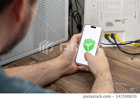 A person holds a smartphone displaying a green shield icon to demonstrate secure home network settings and effective wireless internet protection in a modern digital workspace area A person holds a smartphone displaying a green shield icon to demonstrate secure home network settings and effective wireless internet protection in a modern digital workspace area 143500650