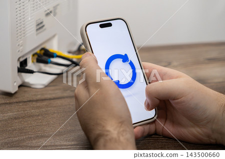 A person holds a smartphone displaying a refresh icon next to a home wireless internet router, representing network connectivity, technical support, and managing digital service flow A person holds a smartphone displaying a refresh icon next to a home wireless internet router, representing network connectivity, technical support, and managing digital service flow 143500660