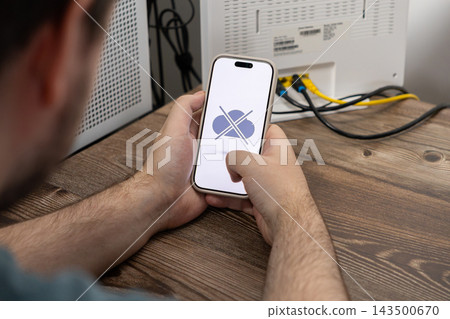 Person holds a smartphone showing a cloud connection error icon while troubleshooting a wireless internet router at a wooden desk to illustrate technical issues and network outages Person holds a smartphone showing a cloud connection error icon while troubleshooting a wireless internet router at a wooden desk to illustrate technical issues and network outages 143500670