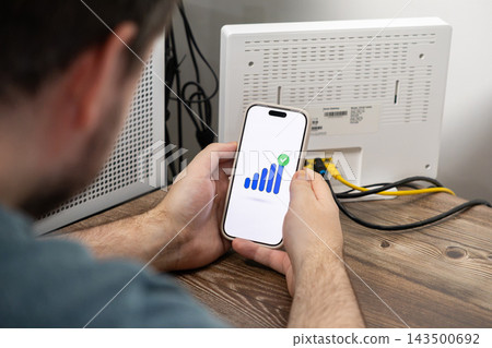 Person holding a smartphone displaying a signal strength icon while standing beside a wireless internet router, representing successful network connectivity and home technology setup Person holding a smartphone displaying a signal strength icon while standing beside a wireless internet router, representing successful network connectivity and home technology setup 143500692