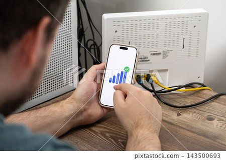 A person checks their strong wifi signal strength on a smartphone app screen next to a home router to ensure reliable network connectivity for remote work and digital management A person checks their strong wifi signal strength on a smartphone app screen next to a home router to ensure reliable network connectivity for remote work and digital management 143500693