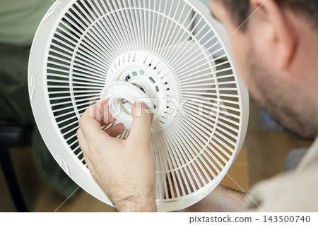 Technician assembling a white electric fan, attaching the plastic nut to secure the safety grille and blade cover during home appliance maintenance in a bright living environment room Technician assembling a white electric fan, attaching the plastic nut to secure the safety grille and blade cover during home appliance maintenance in a bright living environment room 143500740