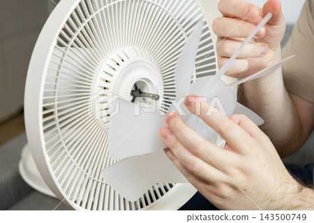 Hands assembling a white plastic electric fan blade onto the motor shaft to restore home cooling device functionality, representing maintenance, repair, and indoor comfort solutions Hands assembling a white plastic electric fan blade onto the motor shaft to restore home cooling device functionality, representing maintenance, repair, and indoor comfort solutions 143500749
