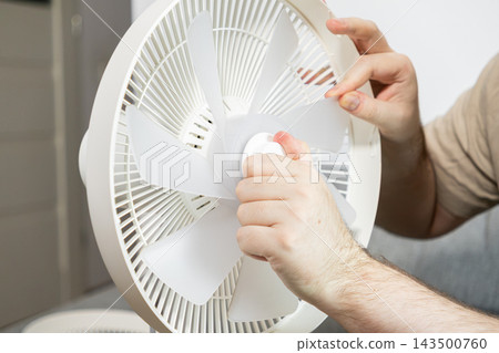 Person assembling a white portable electric fan by securing plastic blades to the motor housing, highlighting routine home maintenance and appliance repair tasks in a modern residence Person assembling a white portable electric fan by securing plastic blades to the motor housing, highlighting routine home maintenance and appliance repair tasks in a modern residence 143500760