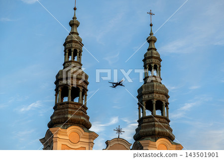 A passenger airplane flies between twin stone church towers under a clear blue sky, illustrating the juxtaposition of religious architecture and modern aviation travel in urban space A passenger airplane flies between twin stone church towers under a clear blue sky, illustrating the juxtaposition of religious architecture and modern aviation travel in urban space 143501901