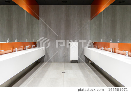 Modern public restroom interior with long white twin washbasins, gray tile walls, and vibrant orange glass accents, creating a professional atmosphere in a clean contemporary facility Modern public restroom interior with long white twin washbasins, gray tile walls, and vibrant orange glass accents, creating a professional atmosphere in a clean contemporary facility 143501971