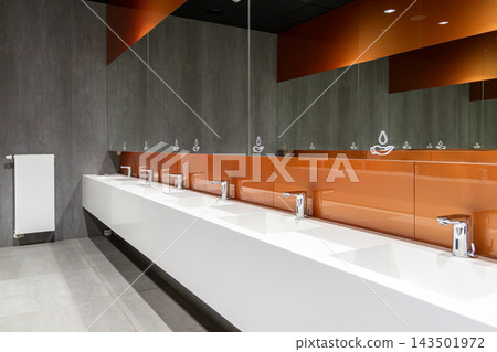Modern public restroom interior with long white washbasin vanity and orange glass accents providing a sleek hygienic environment for corporate building sanitation and facility design Modern public restroom interior with long white washbasin vanity and orange glass accents providing a sleek hygienic environment for corporate building sanitation and facility design 143501972