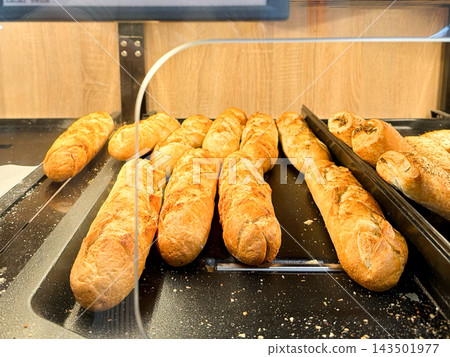 Fresh artisan baguettes arranged on a black metal bakery tray behind a glass display case, conveying a warm morning atmosphere for a local grocery store or a neighborhood cafe shop Fresh artisan baguettes arranged on a black metal bakery tray behind a glass display case, conveying a warm morning atmosphere for a local grocery store or a neighborhood cafe shop 143501977
