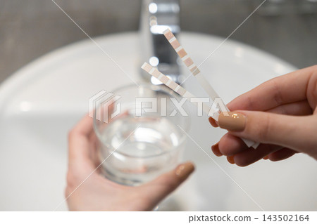 A hand holds liquid testing strips over a glass of water to check quality and safety, representing home analysis of tap purity for health awareness in a bright modern kitchen space A hand holds liquid testing strips over a glass of water to check quality and safety, representing home analysis of tap purity for health awareness in a bright modern kitchen space 143502164