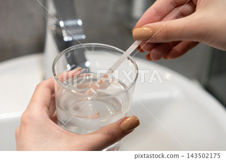 Person testing water quality with a paper strip in a clear glass at a bathroom sink to check pH levels and purity, ensuring safety and cleanliness in a modern domestic environment 143502175