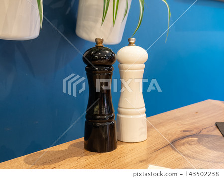 A black and white pepper mill set stands on a rustic wooden table against a blue wall, reflecting minimalist kitchen design and culinary preparation in a modern restaurant space A black and white pepper mill set stands on a rustic wooden table against a blue wall, reflecting minimalist kitchen design and culinary preparation in a modern restaurant space 143502238